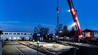 Es früh am Morgen und noch dunkel. Ein Kran und Bauarbeiter arbeiten an einem Umspannwerk.