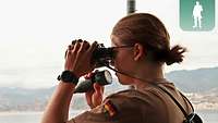 Eine Soldatin steht an Deck und beobachtet mit dem Fernglas das Meer