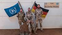 Drei Soldaten stehen vor einem Gebäude, hinter ihnen eine Deutschlandflagge und eine Flagge der VN