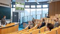 Herr Müller steht am Rednerpult, auf einer Leinwand ist eine Präsentation zu sehen