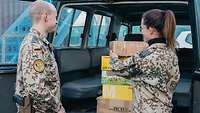 Eine Soldatin und ein Soldat stehen hinter einem Fahrzeug mit geöffneter Heckklappe, im Fahrzeug viele Pakete