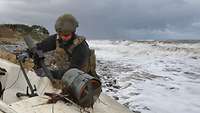 Ein Soldat in blauem Arbeitsanzug, Flecktarnwest und Gefechtshelm hantiert an einem Boot an einem Strand.