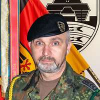 Ein Soldat mit einer goldenen Kordel steht vor einer Truppenfahne.