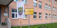Ein zweistöckiges Gebäude: Davor stehen eine Fahne mit dem Bataillonswappen und ein Ortsschild.