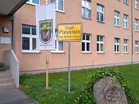 Ein zweistöckiges Gebäude: Davor stehen eine Fahne mit dem Bataillonswappen und ein Ortsschild.