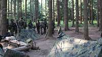 Im Wald stehen zwei Zwei-Mann-Zelte und eine Feuerstelle, im Hintergrund Soldaten.
