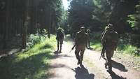 Mehrere Soldaten laufen einen Waldweg entlang, in Abständen, links und rechts aufgeteilt. 
