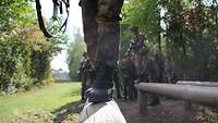 Ein Soldat balanciert über einen Holzbalken. Im Hintergrund warten Soldaten, bis sie dran sind.