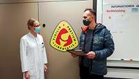 Ein Mann hält einen Bilderrahmen in der Hand. Neben ihm steht eine Frau in weißen Kittel und sieht ihn an.