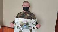 Ein Soldat hält einen Bilderrahmen in den Händen, in denen eine Collage aus Postkarten zu sehen ist