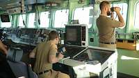 Auf einem Schiff sieht ein Soldat durch ein Fernglas. Im Hintergrund sitzt ein Soldat an einem Tisch mit Kartenausschnitten.