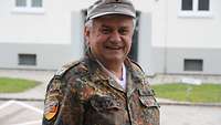 Ein Soldat im Feldanzug und mit Bergmütze steht vor eine Gebäude und lächelt in die Kamera.