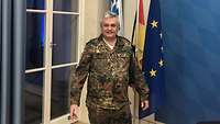 Ein Soldat, gut gelaunt, steht im Feldanzug vor drei Fahnen und einer Wand mit blauem Rautenmuster. 