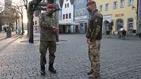 Ein deutscher und ein ungarischer Soldat stehen auf einem gepflasterten Platz, im Hintergrund eine Häuserzeile mit Geschäften. 