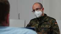 Oberstabsarzt Neumann sitzt mit Mund-Nasen-Schutzmaske im Flecktarnuniform zum Interview an einem Tisch
