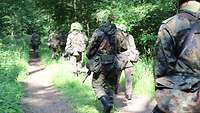 Mehrere getarnte Soldaten laufen auf einem Waldweg hintereinander.