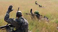 Soldaten hocken und liegen in großen Abständen in hohem Gras und geben sich gegenseitig Handzeichen.