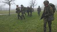 Vier Soldaten tragen auf einer Behelfstrage einen Verwundeten, zwei Soldaten sichern.