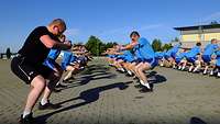 Zahlreiche Soldaten in blauem Sportzeug stehen in Reihen dicht nebeneinander und machen Kniebeugen.