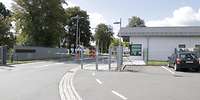 Eine Straße läuft auf den Kaserneneingang mit zwei Schranken zu, rechts ein weißes Wachgebäude.