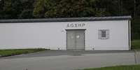 Helles, flaches Gebäude ohne Fenster vor einem Waldstück, über dem Eingang die Buchstaben AGSHP.
