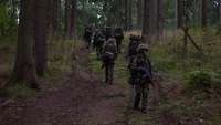 Eine Gruppe von Soldaten marschiert mit Gepäck eine Anhöhe im Wald hinauf, ein Soldat dreht sich um.