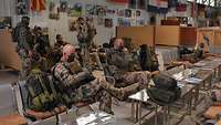 Soldaten sitzen und stehen in einer Halle, an den Wänden hängen Flaggen verschiedener Nationen
