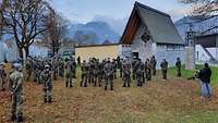 Zahlreiche Soldaten stehen vor einer kleinen, steinernen Kapelle. Im Hintergrund erheben sich Berge.