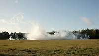 A dense smoke screen on a green field in front of woods