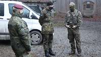 Drei Soldaten stehen auf einem Parkplatz an einem Waldrand und sprechen miteinander.