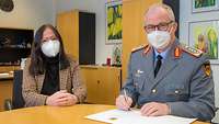 Männliche Person in Uniform schreibt in ein Heft auf einem Tisch, neben ihm sitz weibliche Person in zivil auf Abstand