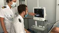 Ein Mann sitzt vor einem Monitor und hält ein medizinisches Handgerät an den Bauch eines liegenden Patienten