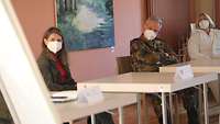 Drei Personen sitzen hinter jeweils einem Tisch mit Abstand zueinander in einem Vortragsraum