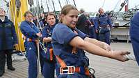 Drei Marinesoldatinnen in blauen Overalls stehen auf dem Oberdeck eines Segelschiffs und ziehen an einer Leine.