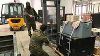 Ein Soldat verzurrt Kisten auf einer Palette, während ein Kamerad mit einem Hubwagen unter die Palette fährt