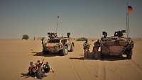 Zwei Spähwagen Fennek stehen in der Wüste, im Vordergrund spielen einheimische Kinder im Sand