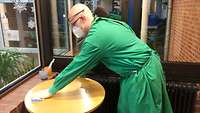 Feldwebel Dennis Lemmen wischt einen runden Tisch ab. Dabei trägt er unter anderem einen Schutzmantel, eine Maske und Handschuhe