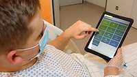 Ein männlicher Patient liegt im Krankenhausbett und schaut sich ein Tablet an