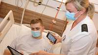 Eine Krankenschwester hält ein Smartphone in der rechten Hand und erklärt einem männlichen Patienten den Umgang mit dem Tablet.
