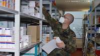 Ein Soldat mit Gesichtsschutz steht an einem Regal mit medizinischen Produkten und hält eine Liste in der Hand.