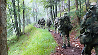 Mehrere Soldaten marschieren mit Gepäck und Waffen hintereinander einen Waldweg im Gebirge entlang.