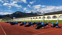 Soldaten in blauer Sportbekleidung führen auf einer Tartanbahn gleichzeitig Fitnessübungen aus.