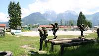 Zwei Soldaten mit Marschgepäck und Waffen balancieren über runde Holzbalken.