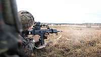 Ein Soldat liegt mit Helm und einer Waffe in einer Stellung und schaut auf das vor ihm liegende Feld.