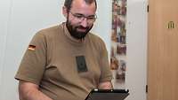 Ein Soldat mit einem Tablet in der Hand in seinem Zimmer, im Hintergrund hängen Bilder seiner Familie an der Wand 