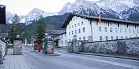 Eine Straße läuft auf eine Einfahrt mit Schlagbaum zu, dahinter ein dreistöckiges, helles Gebäude.