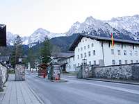 Eine Straße läuft auf eine Einfahrt mit Schlagbaum zu, dahinter ein dreistöckiges, helles Gebäude.