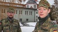 Ein Soldat und eine Soldatin schauen freundlich in die Kamera, hinter ihnen ein grau-weißes Gebäude und Schnee.