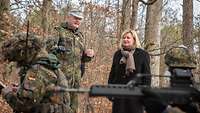Eine Zivilistin steht im Wald neben einem Soldaten, der ihr etwas erklärt. Ein Soldat hält ein Gewehr.