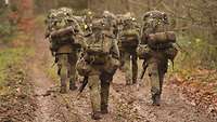 Mehrere Soldaten marschieren mit großen Rucksäcken einen Waldweg entlang.